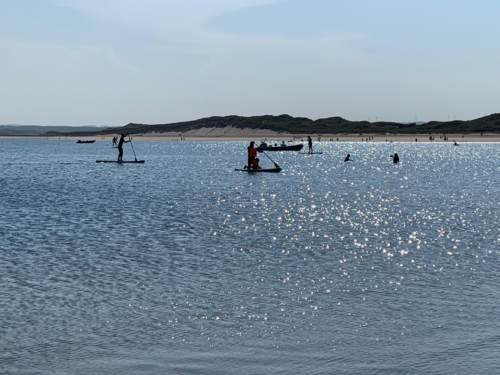 Beadnell Bay beach. People in the sea and on the beach. Stand Up paddle-boarders are in the foreground. The weather looks hot.