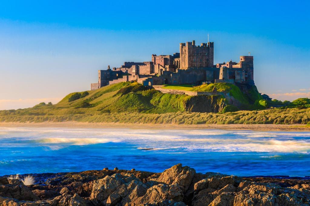 Photo of the imposing Bamburgh Castle, with rocks in the foreground. The photo has been edited so that the sea is cerulean blue, and the sky a vivid blue also